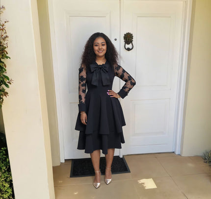 Black lace-sleeve layered midi dress with bow neckline and tiered skirt.