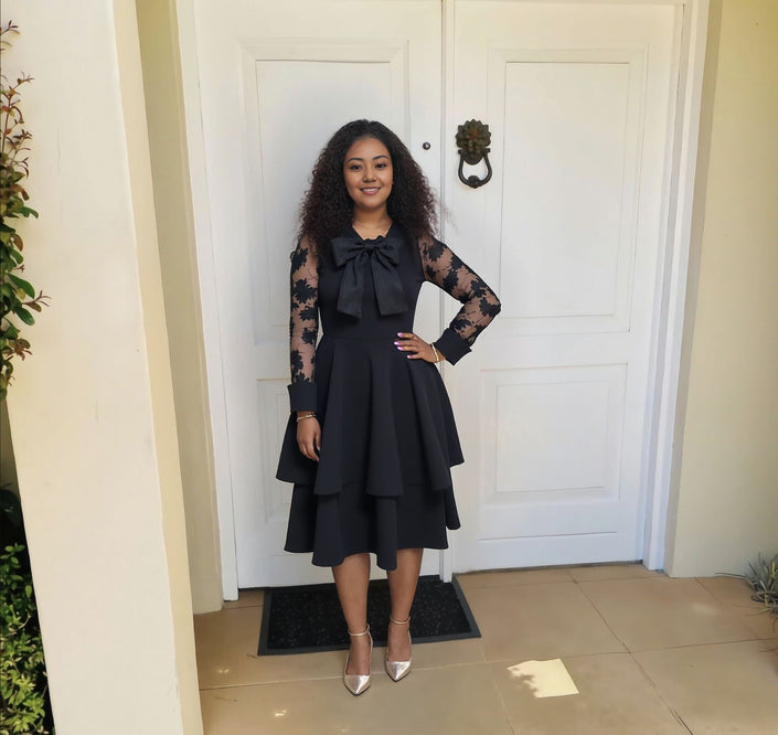 Black lace-sleeve layered midi dress with bow neckline and tiered skirt.